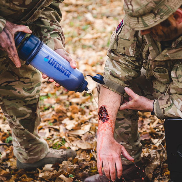 Load image into Gallery viewer, Person in camouflage holding a blue 'Lifesaver' Bottle over another person's wound outdoors.