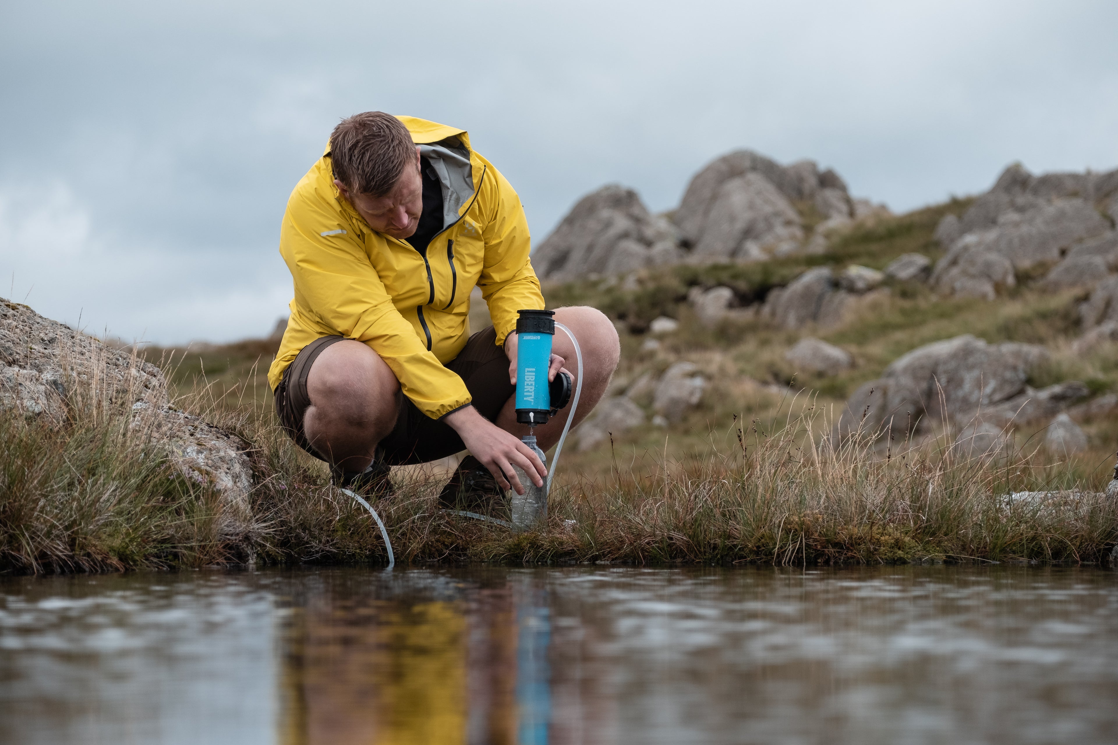 Person in a yellow jacket collecting water from a stream in a rocky landscape