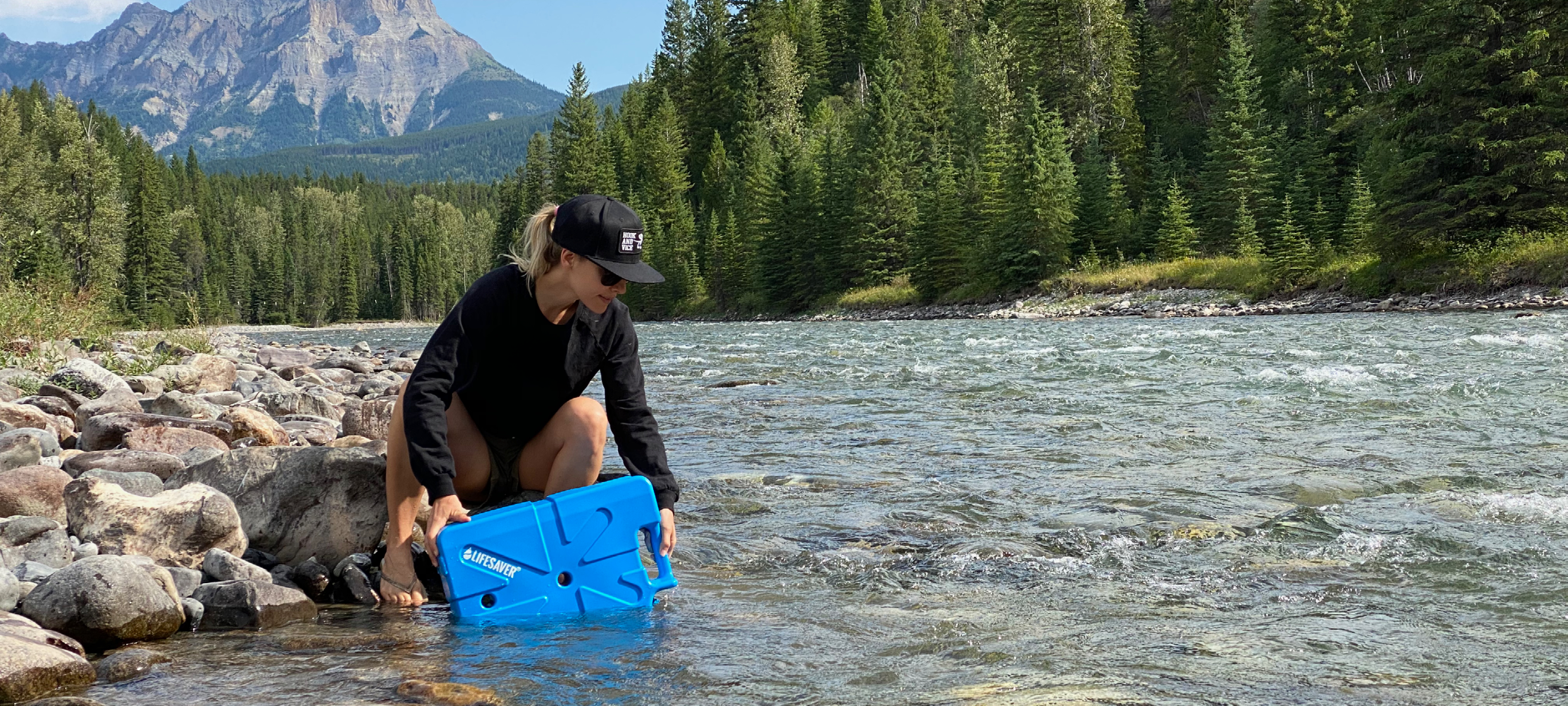 Woman filling a blue LifeSaver Jerrycn from a river with trees and mountains in the background