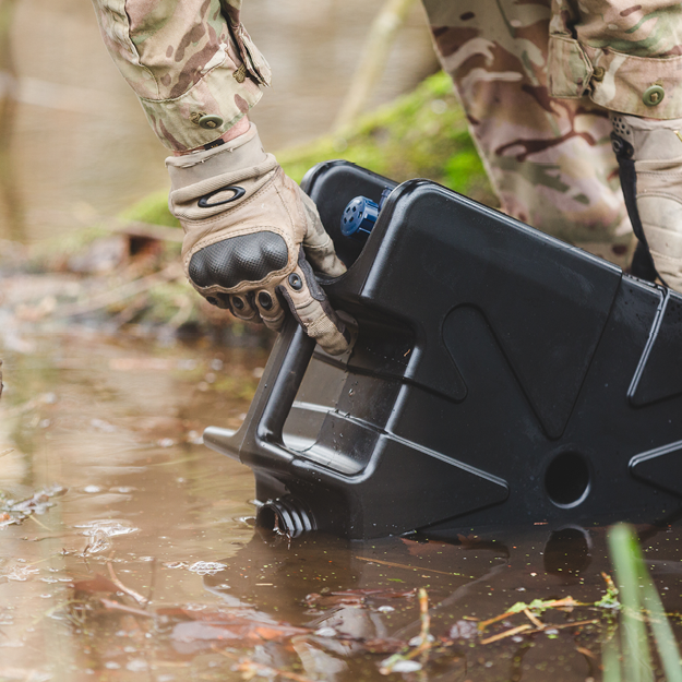 Person in camouflage gear holding a black jerry can in a muddy outdoor setting