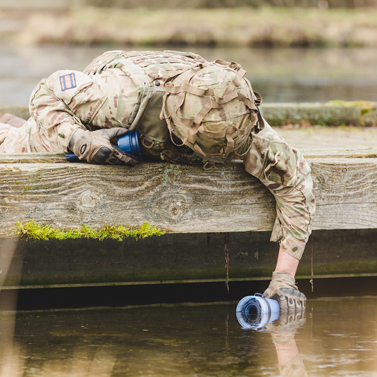 Person in military camouflage lying on a wooden dock with a LifeSaver Bottle, surrounded by water and greenery.